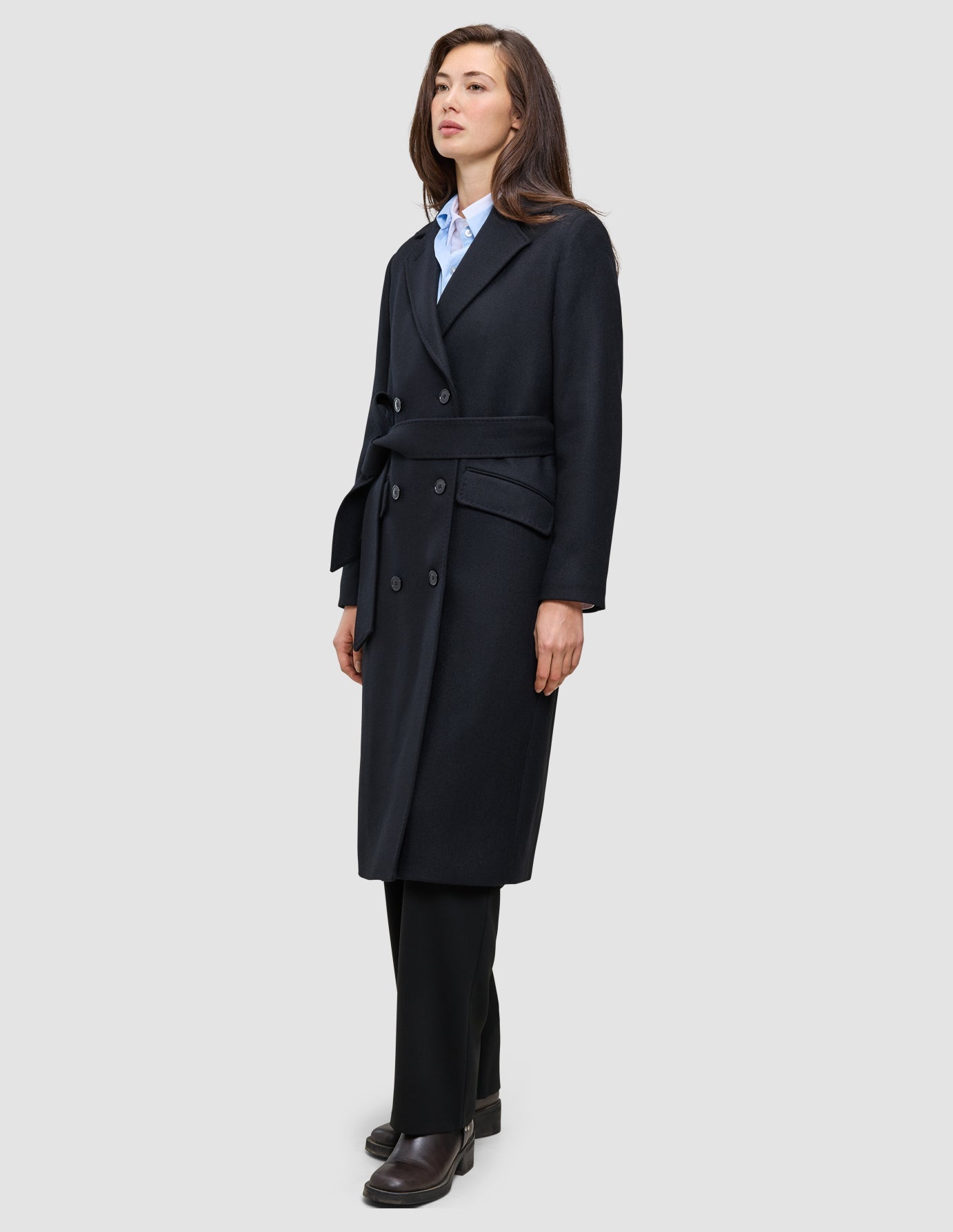 A woman wearing a Women’s Wool Coat Black in a Scandinavian-design double-breasted style with a tie waist over a light blue shirt and black trousers.